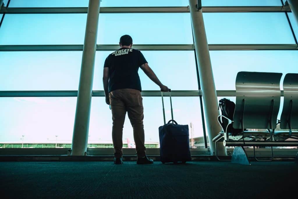 Luggage Buying Mistakes: What Travelers Often Overlook 5 Man standing inside an airport terminal with a carry-on suitcase, looking out the large glass windows before his flight.