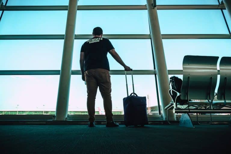 Man standing inside an airport terminal with a carry-on suitcase, looking out the large glass windows before his flight.