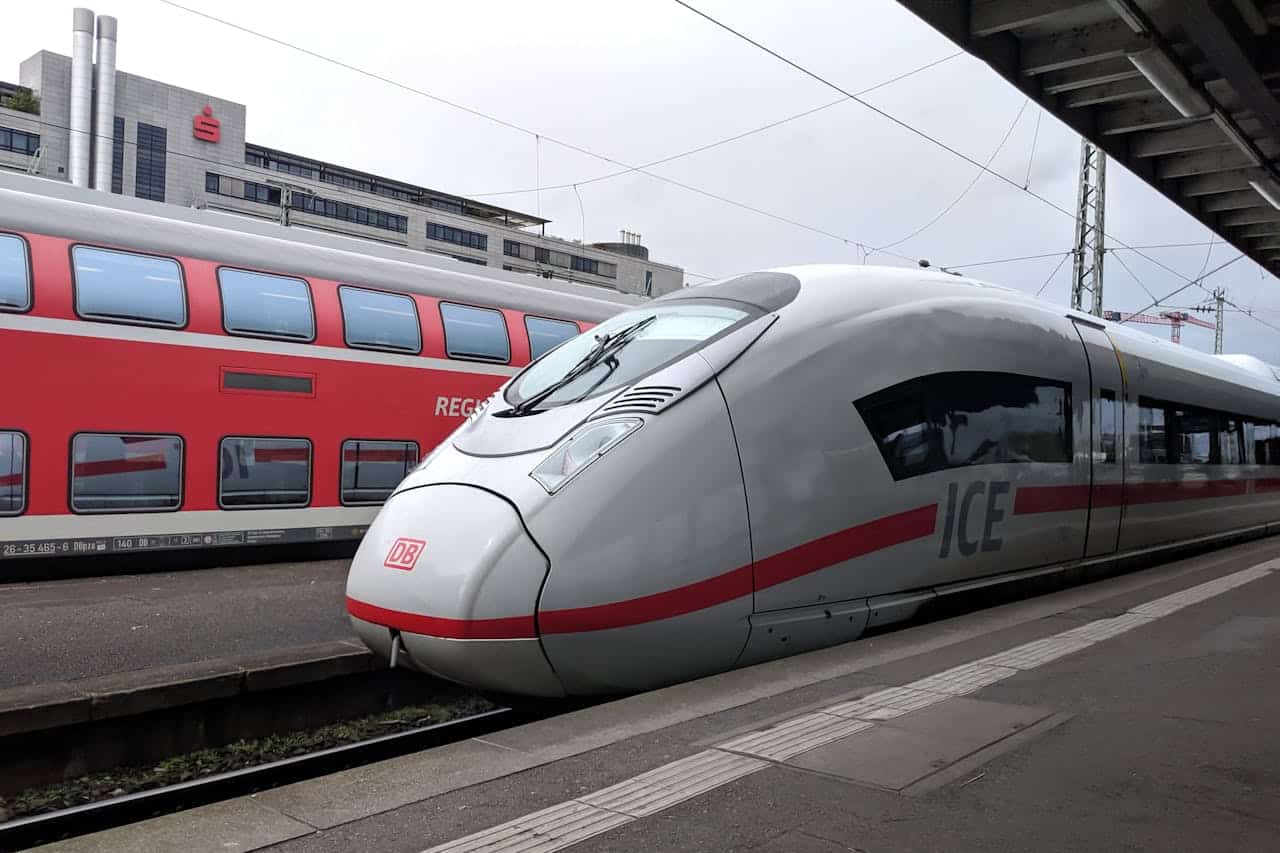 A modern German ICE high-speed train at a station platform, with a red regional train in the background.