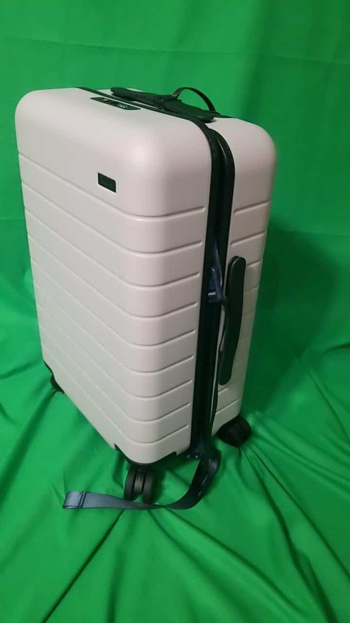 Away Carry-On suitcase in beige standing upright with zipper slightly open against a green backdrop.