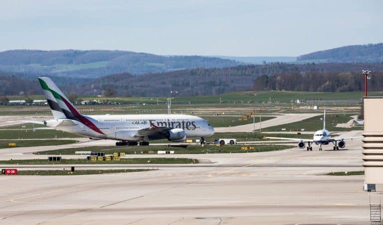 Emirates Airbus A380 taxiing on the runway with another aircraft approaching in the background at an international airport surrounded by hills.