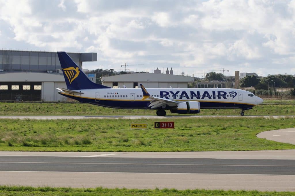 Ryanair Boeing 737 aircraft taxiing on the runway at an airport, with hangars and cloudy sky in the background.