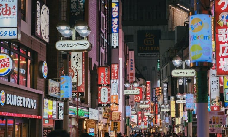 Night street scene in Shibuya, Tokyo filled with bright neon signs, restaurants, and crowds of people walking through the busy shopping district.