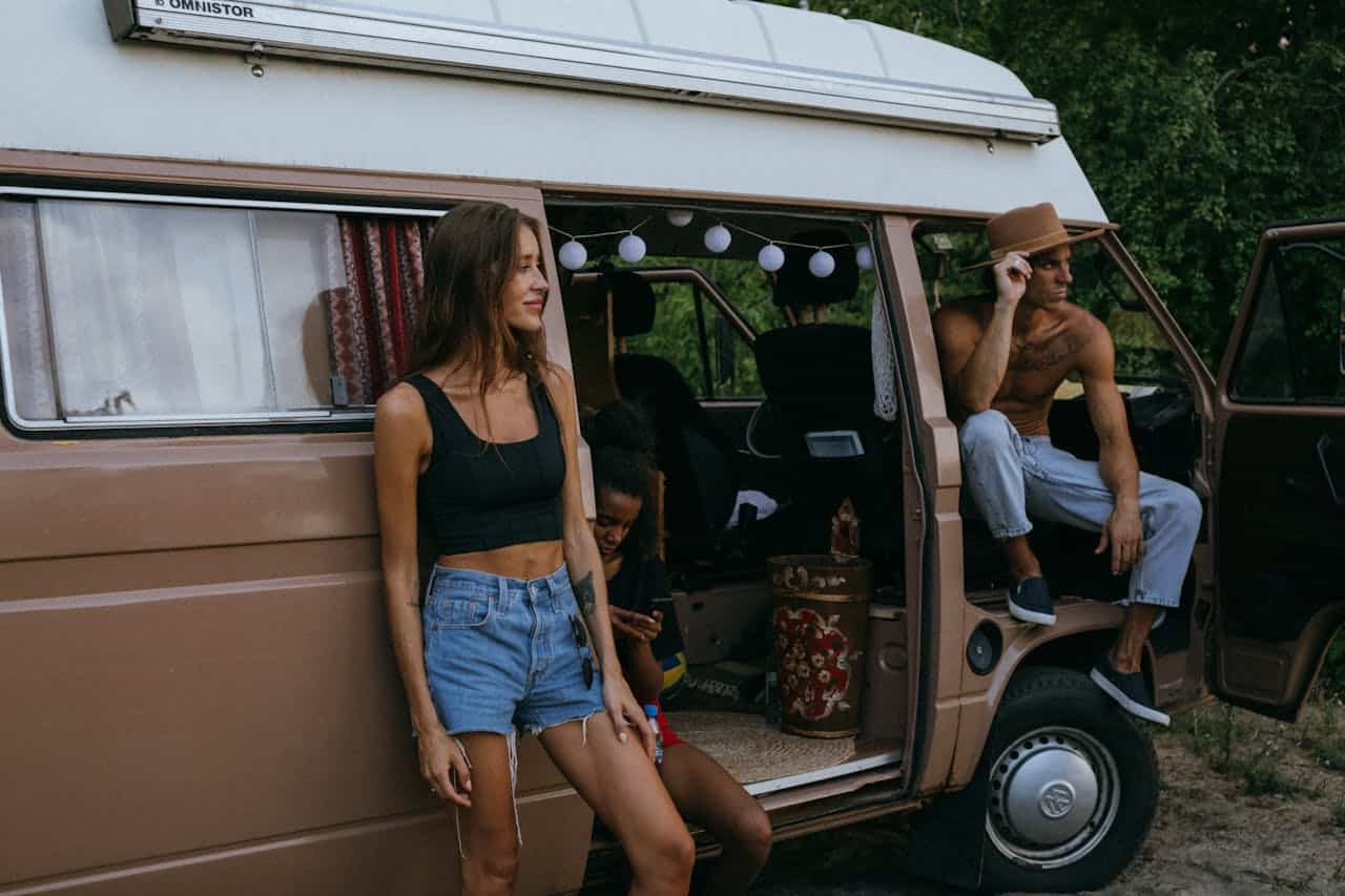 Three people relaxing beside a vintage camper van with the sliding door open, enjoying an outdoor RV stop in a natural setting.