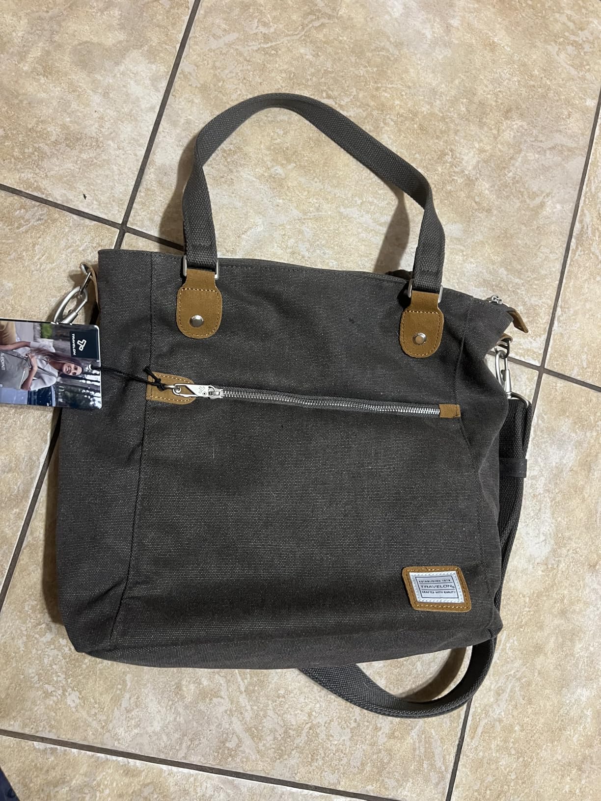 Travelon Anti-Theft Heritage Tote Bag in pewter cotton canvas with suede trim, front zip pocket, and dual handles displayed on tiled floor