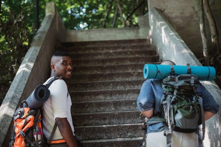 Two men carrying weekend backpacks with rolled sleeping mats hiking up stone stairs in a forest setting