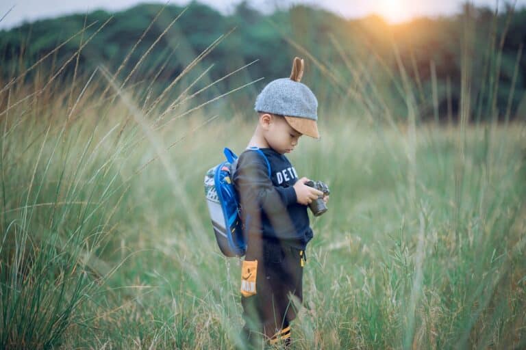 Toddler boy wearing a small blue mini backpack standing in tall grass outdoors during sunset