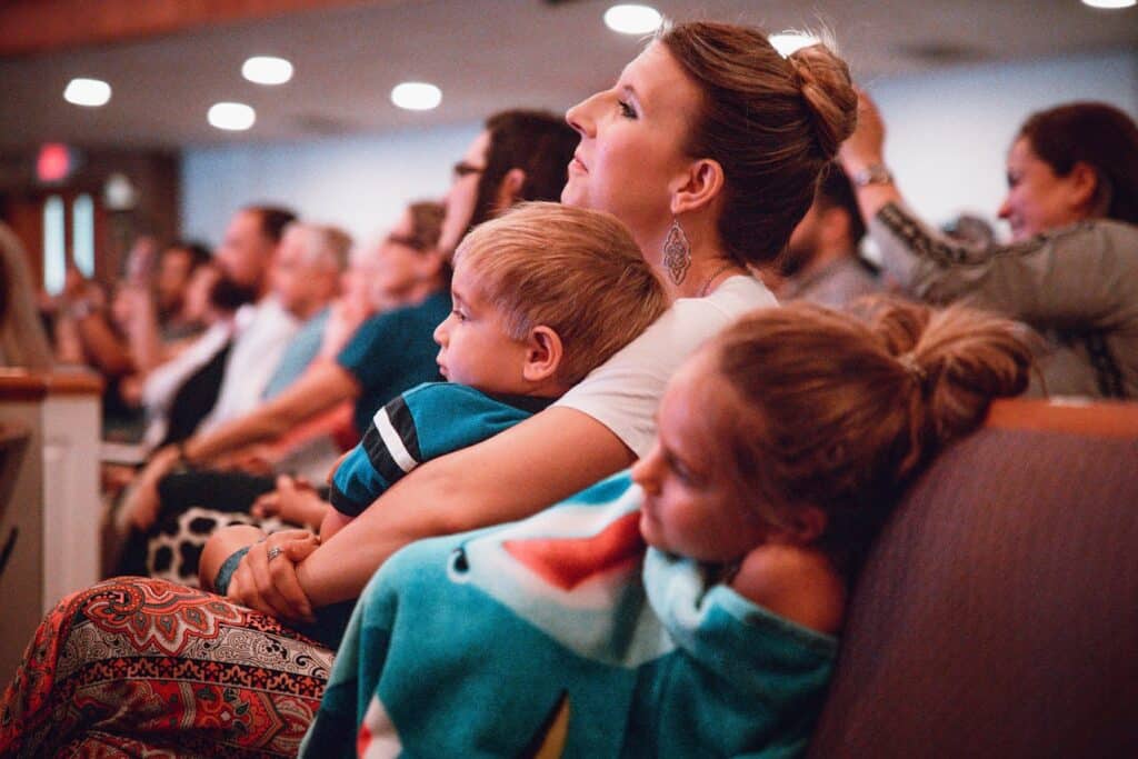 A mother sitting in an auditorium holding her toddler while another young child rests beside her, illustrating the realities of parenting with young children during public events.