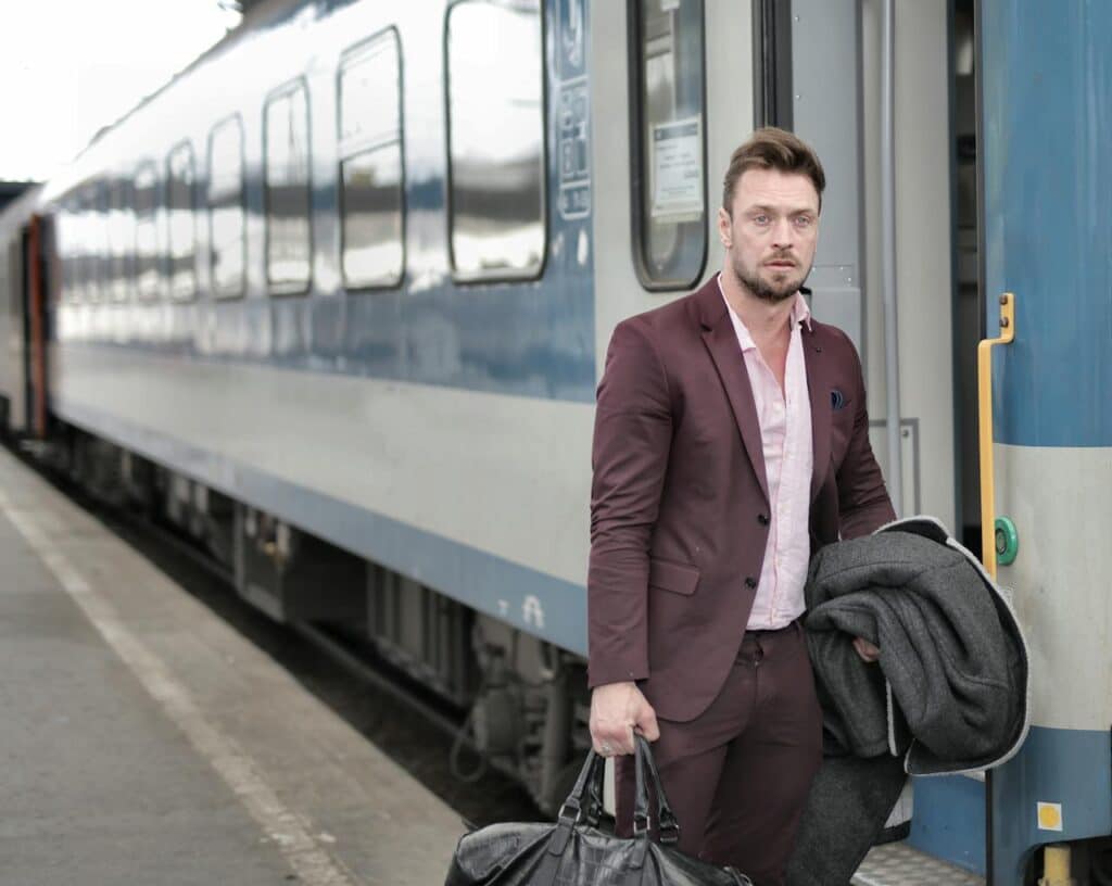 man in maroon suit stepping off train holding leather duffel bag and coat at station platform