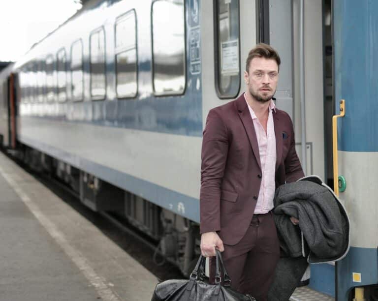 man in maroon suit stepping off train holding leather duffel bag and coat at station platform