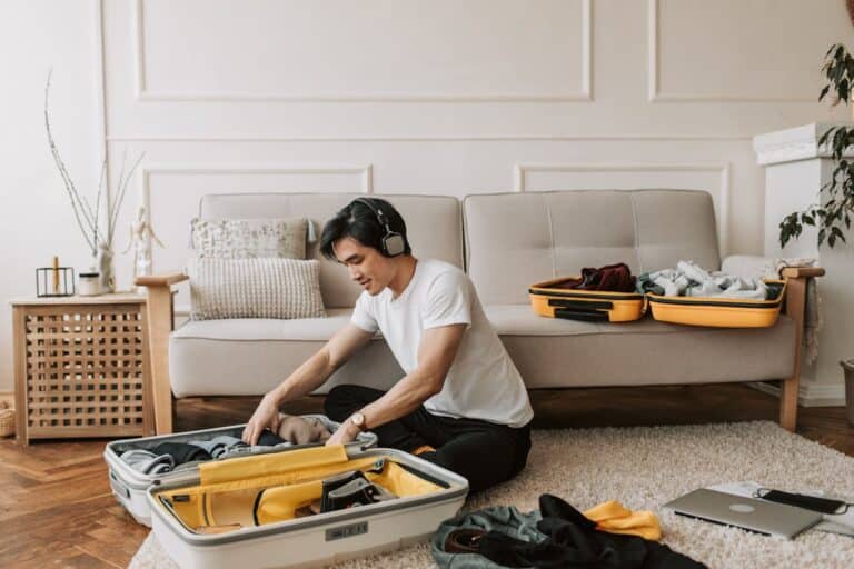 Man packing clothes into an open suitcase on the floor at home, with headphones on and travel items nearby