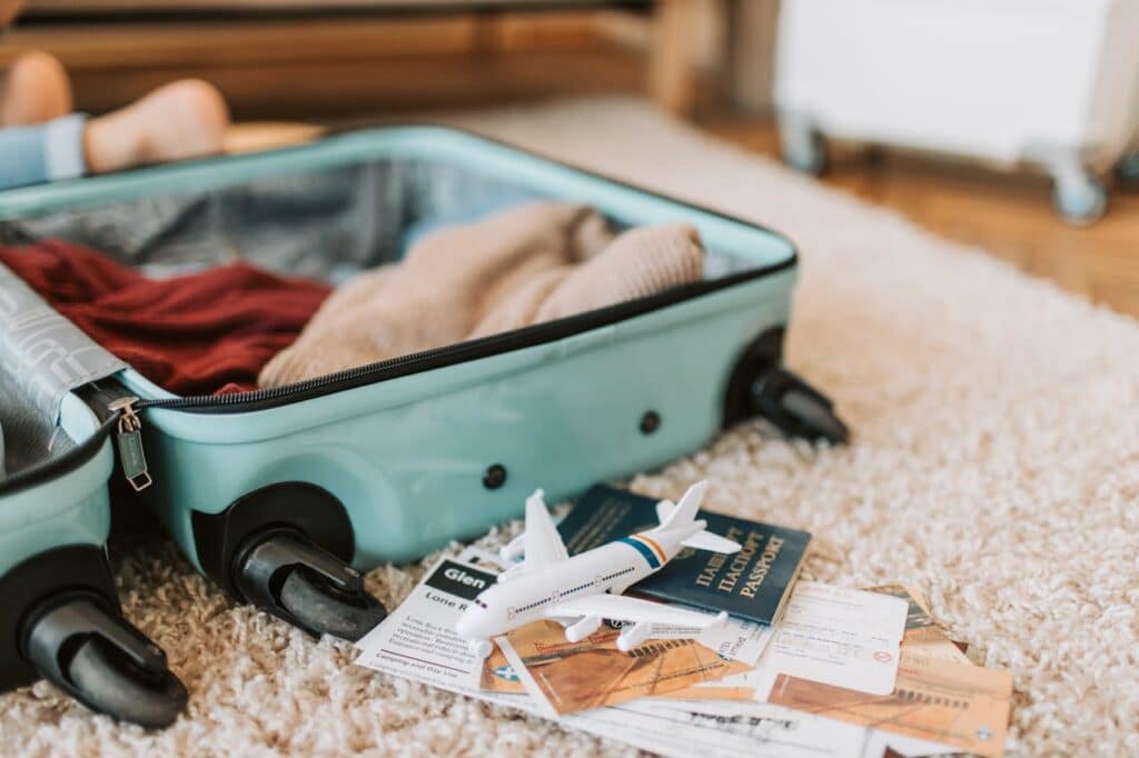 Open mint green cabin suitcase packed with clothes on carpet beside passport, toy airplane, tickets, and travel documents