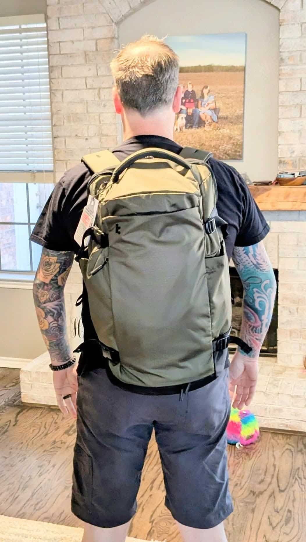 Man wearing an olive green tomtoc travel backpack with padded straps and multiple compartments while standing indoors