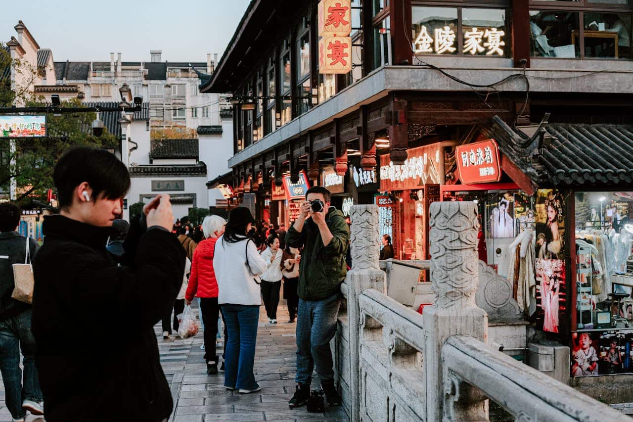 Crowded street in China with tourists taking photos near traditional shops and illuminated signage