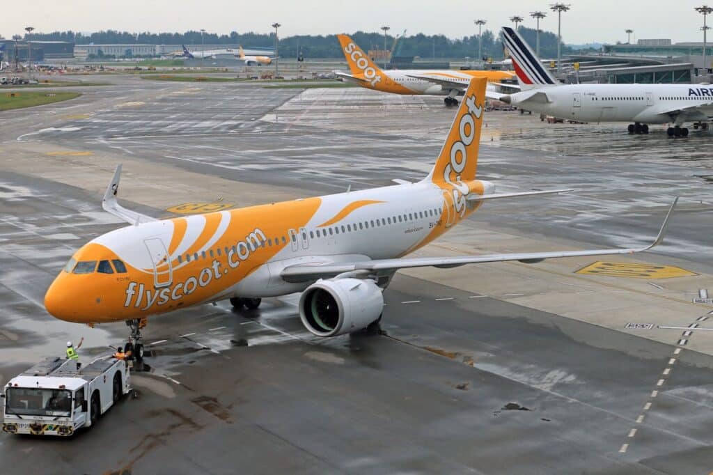 Scoot airplane on airport tarmac preparing for departure with ground crew and terminal in the background