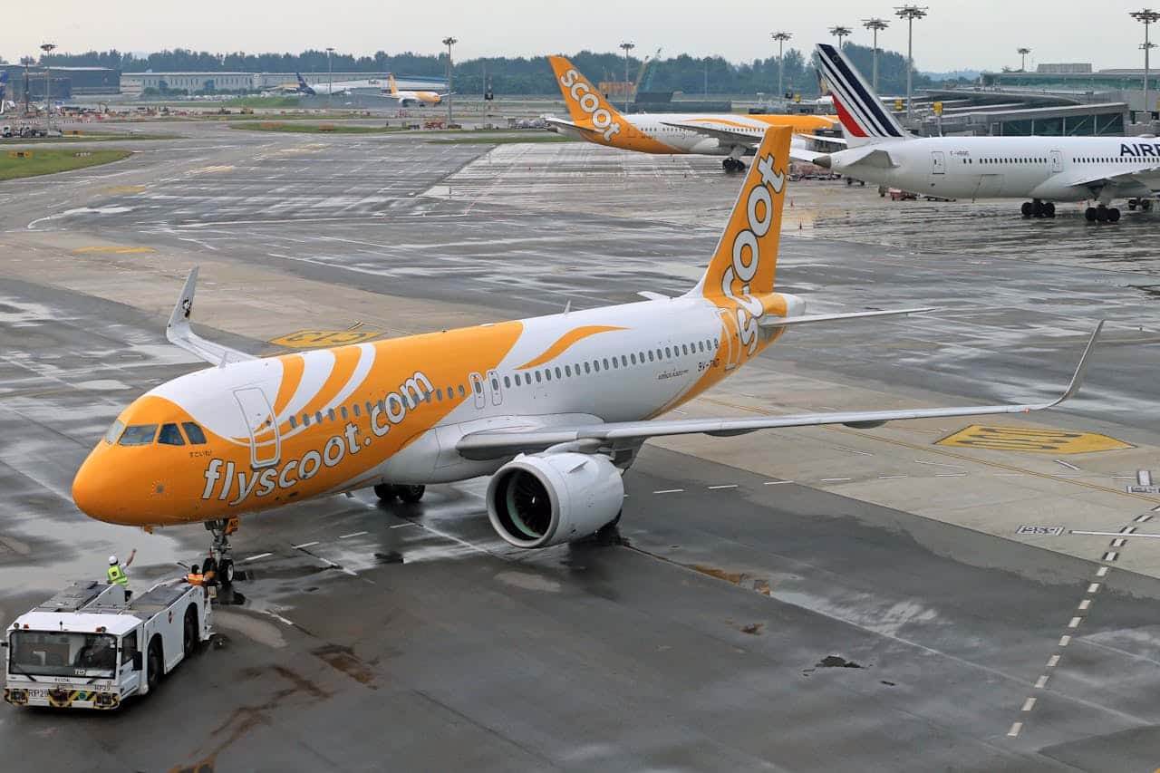 Scoot airplane on airport tarmac preparing for departure with ground crew and terminal in the background