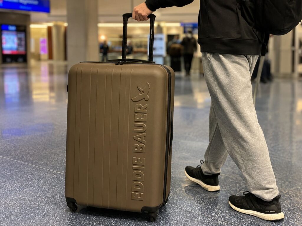 Eddie Bauer Flatiron Check-In Luggage Review 6 Traveler pulling a brown Eddie Bauer hard shell suitcase through an airport terminal with casual travel outfit and spinner wheels visible