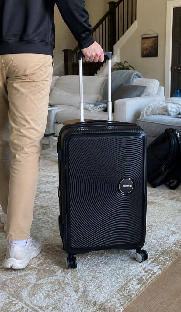 Person pulling a black American Tourister Soundbox suitcase in a living room, lower body visible with casual travel outfit
