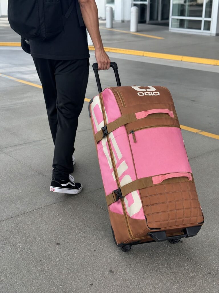 Person pulling OGIO Trucker Roller Gear Bag in pink and brown on a sidewalk, close-up of hand and lower body