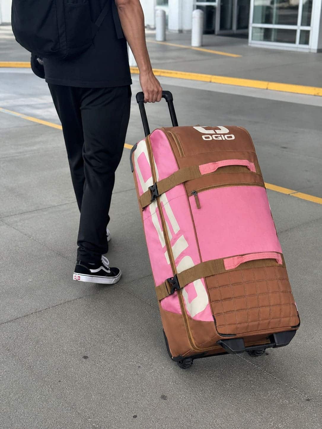 Person pulling OGIO Trucker Roller Gear Bag in pink and brown on a sidewalk, close-up of hand and lower body