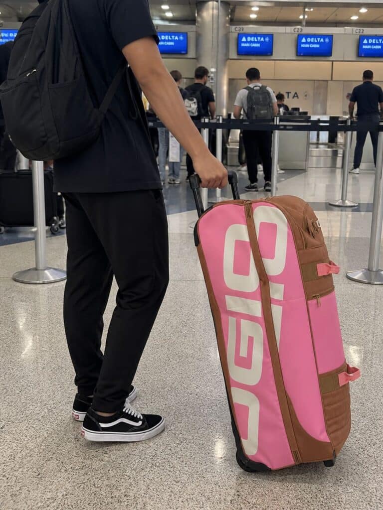 Person standing in airport check-in line holding OGIO Trucker Roller Gear Bag in pink and brown, close-up of hand and lower body