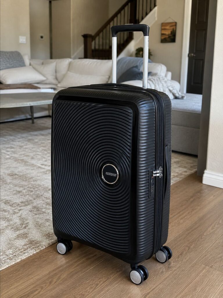 Black American Tourister Soundbox hard-shell suitcase standing in a living room with telescopic handle extended, photographed in natural light