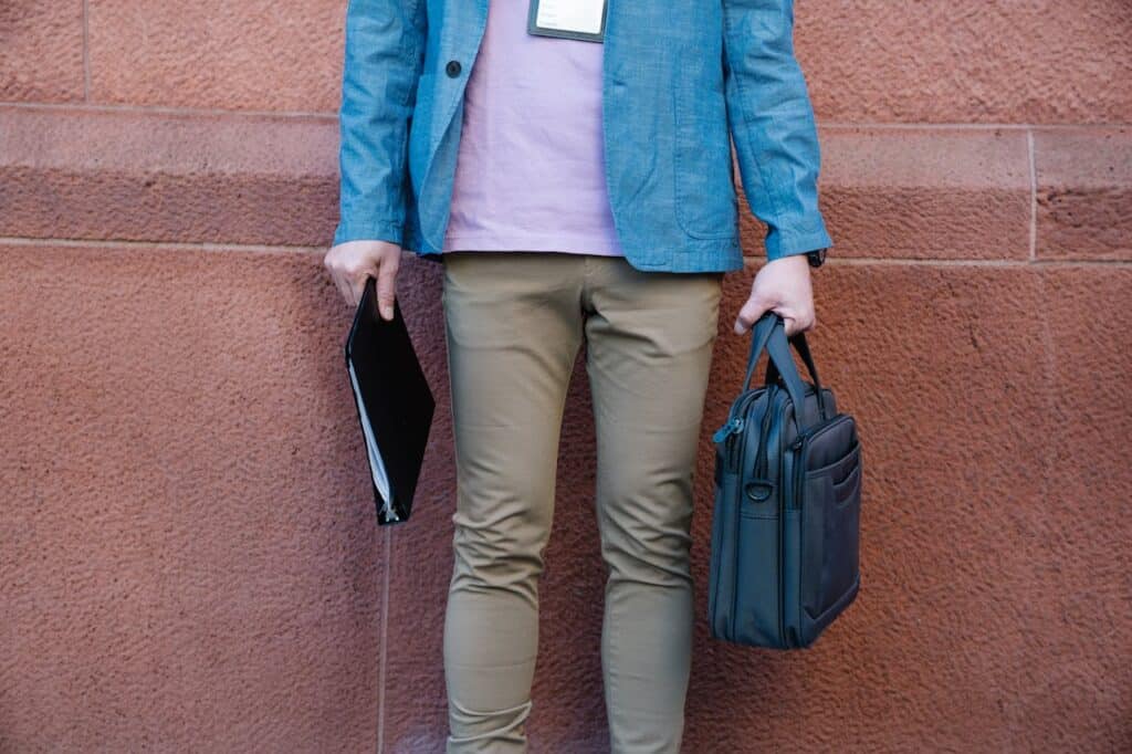 Business traveler holding a slim black laptop bag and document folder while standing against a textured wall outdoors