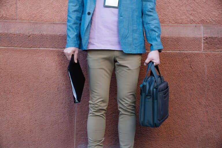 Business traveler holding a slim black laptop bag and document folder while standing against a textured wall outdoors