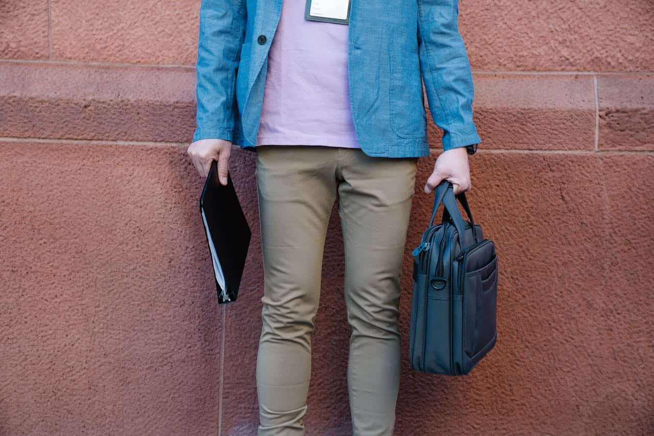 Business traveler holding a slim black laptop bag and document folder while standing against a textured wall outdoors