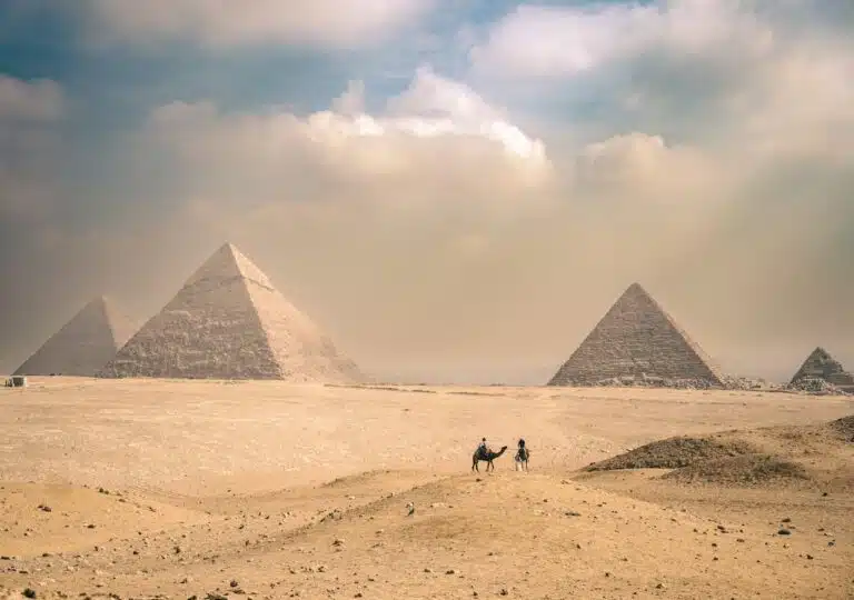 Great Pyramids of Giza in Egypt under a hazy desert sky, with two camel riders crossing the sandy landscape in the foreground.
