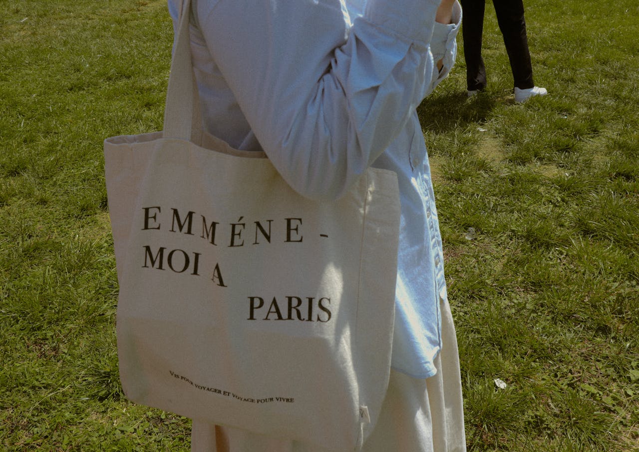 Person carrying a beige canvas tote bag with “Emmène-moi à Paris” text, walking outdoors on grass