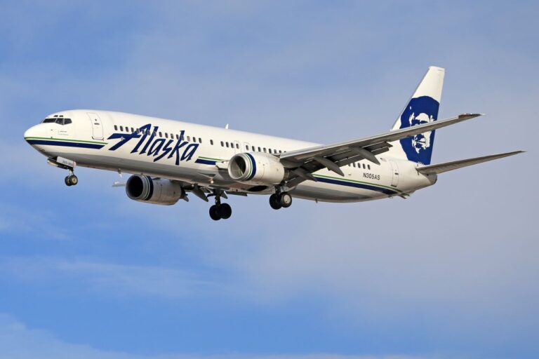Alaska Airlines passenger jet descending with landing gear extended against a blue sky.