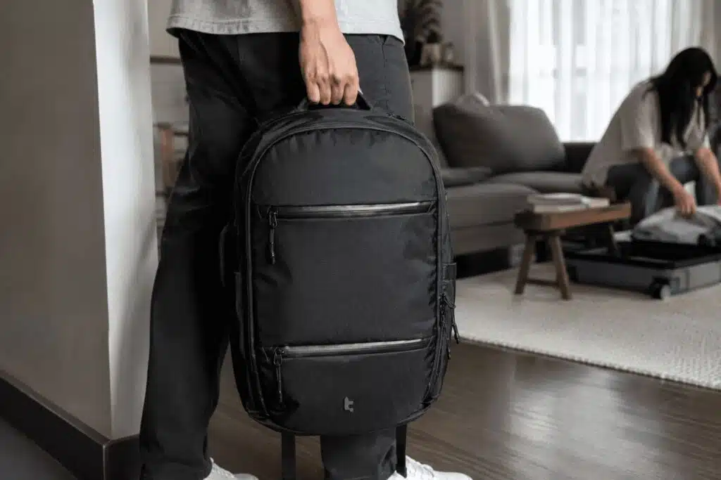 Person holding a black tomtoc UrbanEX-T77 26L laptop backpack while preparing for a trip in a living room.